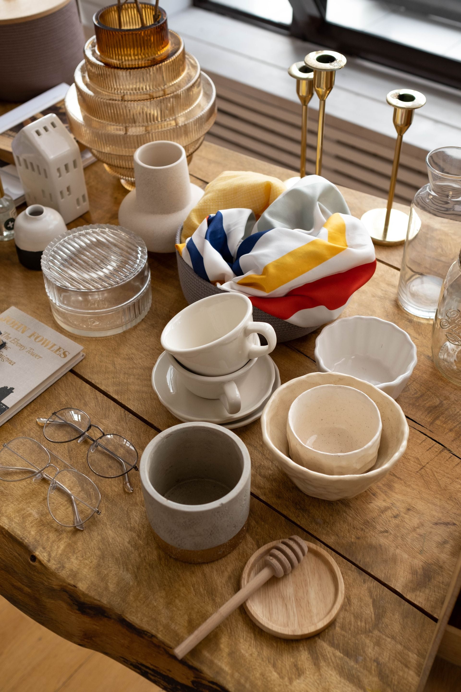 A styled tabletop display of ceramic tableware, including stacked bowls, cups on saucers, a textured gray mug, a wooden honey dipper, and a folded multicolor textile, alongside other home decor items on a rustic wooden table.
