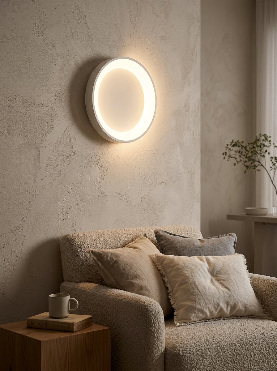 A modern circular LED wall light with a warm white glow mounted on a textured beige plaster wall above a cozy beige sofa with neutral-toned pillows.